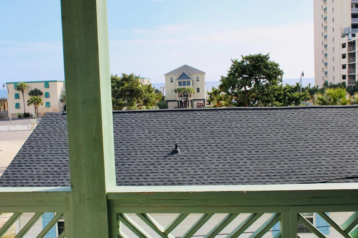 This image captures a tranquil view of the ocean framed by nearby houses and swaying palm trees, with a balcony in the foreground. The atmosphere is serene, highlighting the proximity to the beach and showcasing an inviting spot to relax, watch the sunrise, or enjoy the sea breeze with family and friends.