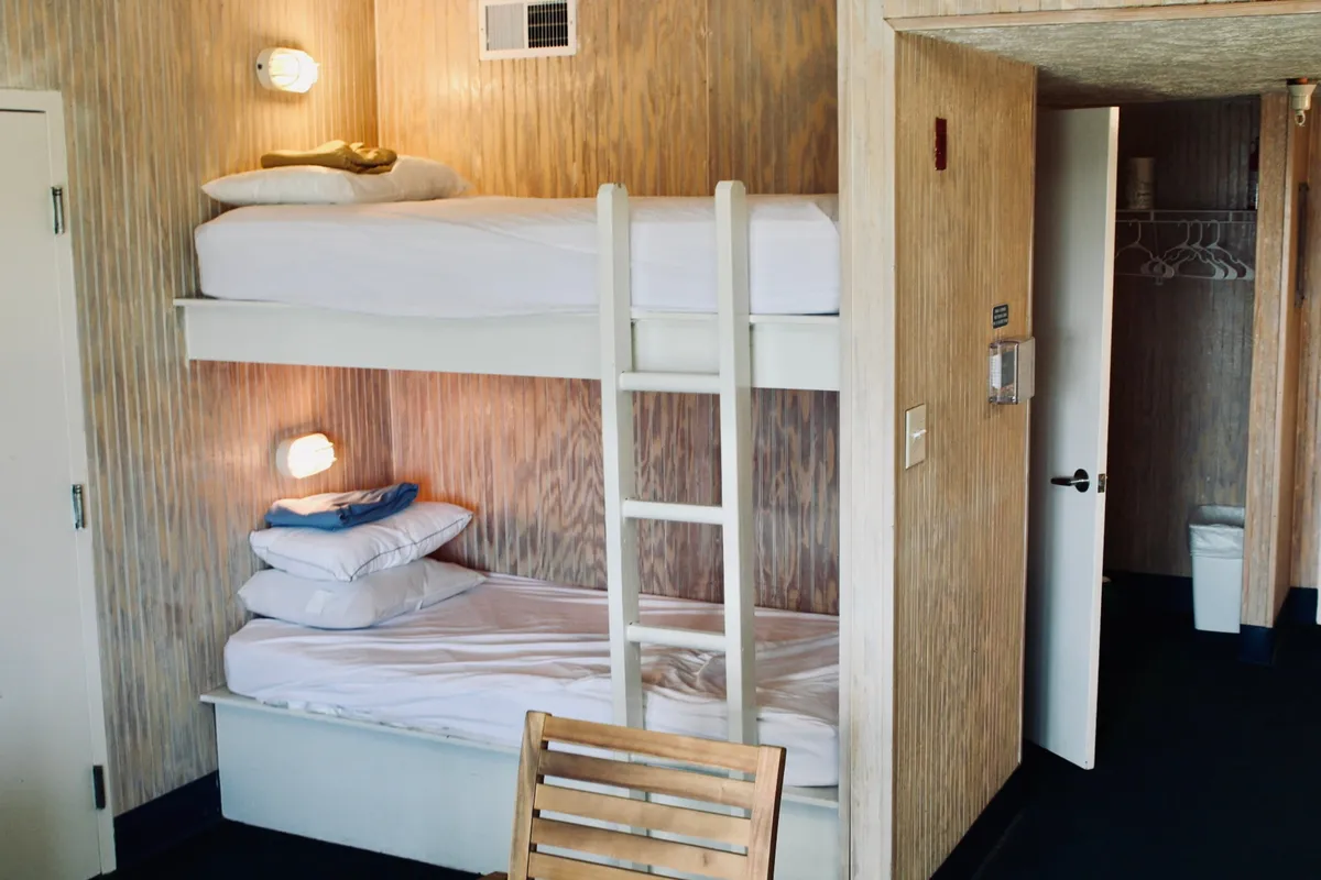 A cozy interior scene featuring bunk beds, a wooden chair, and a doorway leading into another room. The setup evokes a warm, inviting environment suited for casual family routines, morning breakfasts, or winding down after a day out.