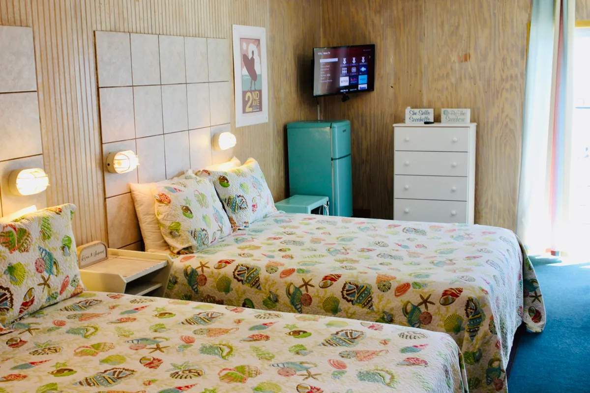 This image features two beds with colorful quilts, a vintage-style refrigerator, a TV mounted on the wall, and a dresser adorned with decorative items. Natural light fills the space, creating a bright and welcoming atmosphere. The arrangement suggests a comfortable area for relaxing, watching movies, sharing meals, or simply enjoying downtime after a day of exploration.