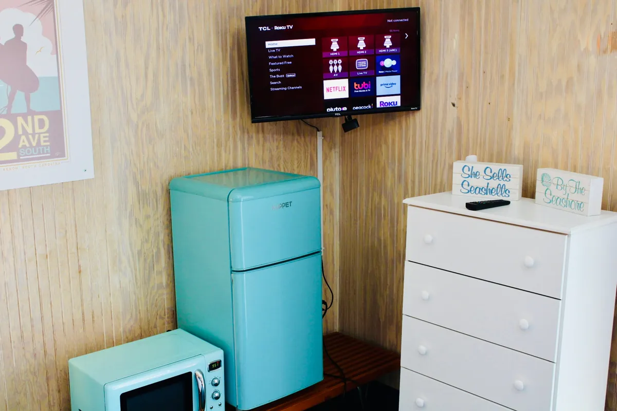 This image showcases a compact kitchenette with a bright turquoise refrigerator, a small microwave perched on a white dresser, a coffeemaker, and subtle decorative touches. The setup is playful yet functional, highlighting spaces for cooking, chilling snacks, and prepping coffee. A TV and streaming options are also visible, adding to the comfort and convenience of the area.